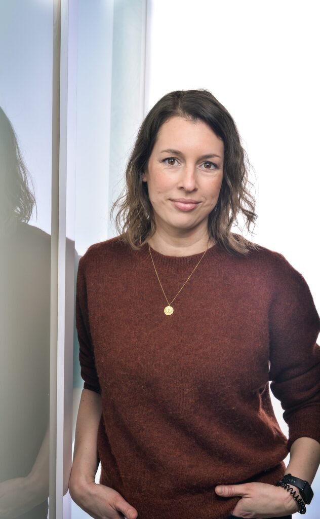 Ein Halbporträt Bild von Evamaria Kalbantner. Sie lehnt an einer Wand. Sie trägt einen rostfarbenen Pullover.